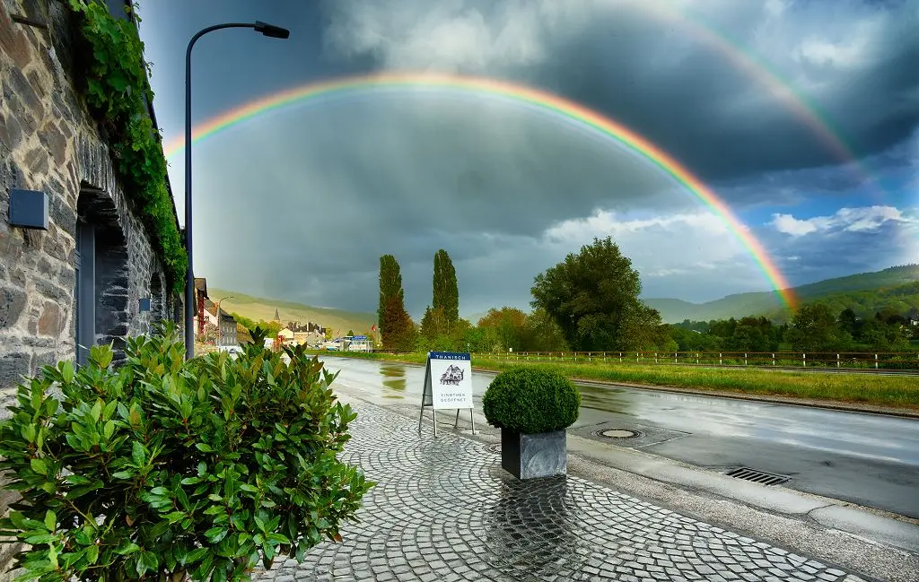 Uitzicht op een dubbele regenboog boven een wijndorp, gezien vanaf een geplaveid trottoir naast een wijnhuis.