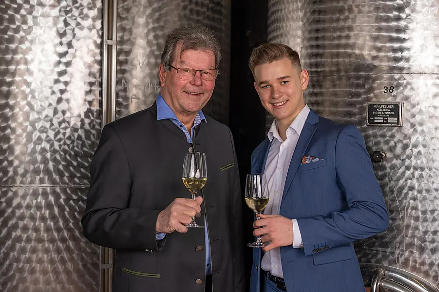 Twee generaties wijnmakers in pak proosten met witte wijn voor grote roestvrijstalen wijnvaten.